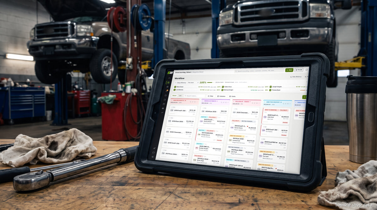 SendWrench workflow board running on a rugged tablet on a tool cart in a diesel repair bay, a lifted truck on a two-post lift behind it